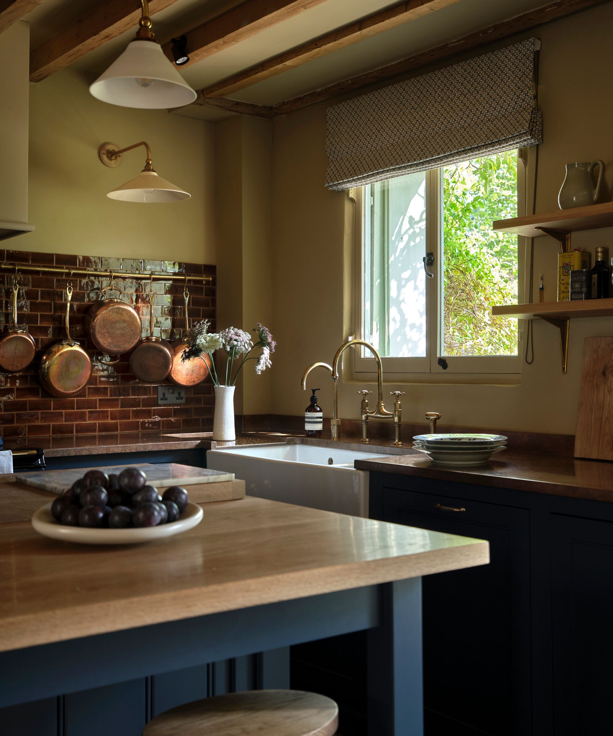 Dark blue kitchen by Violet &amp;amp; George with navy cabinetry, copper backsplash, oak countertops, brass fixtures, exposed beams, and a farmhouse sink.