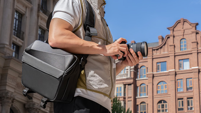 A person stands outdoors, holding a camera in one hand and carrying the MagGoX camera bag over their shoulder, with historic buildings in the background