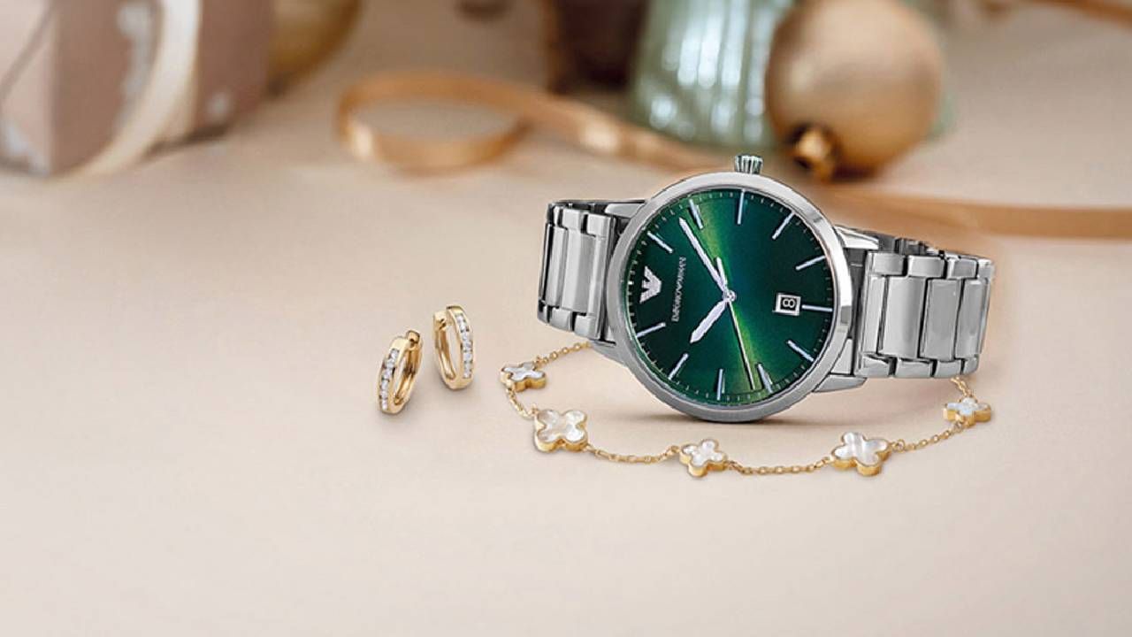 A silver watch with a emerald green watch face with a gold bracelet and a pair of gold earrings from Beaverbrooks in front of a festive backdrop.