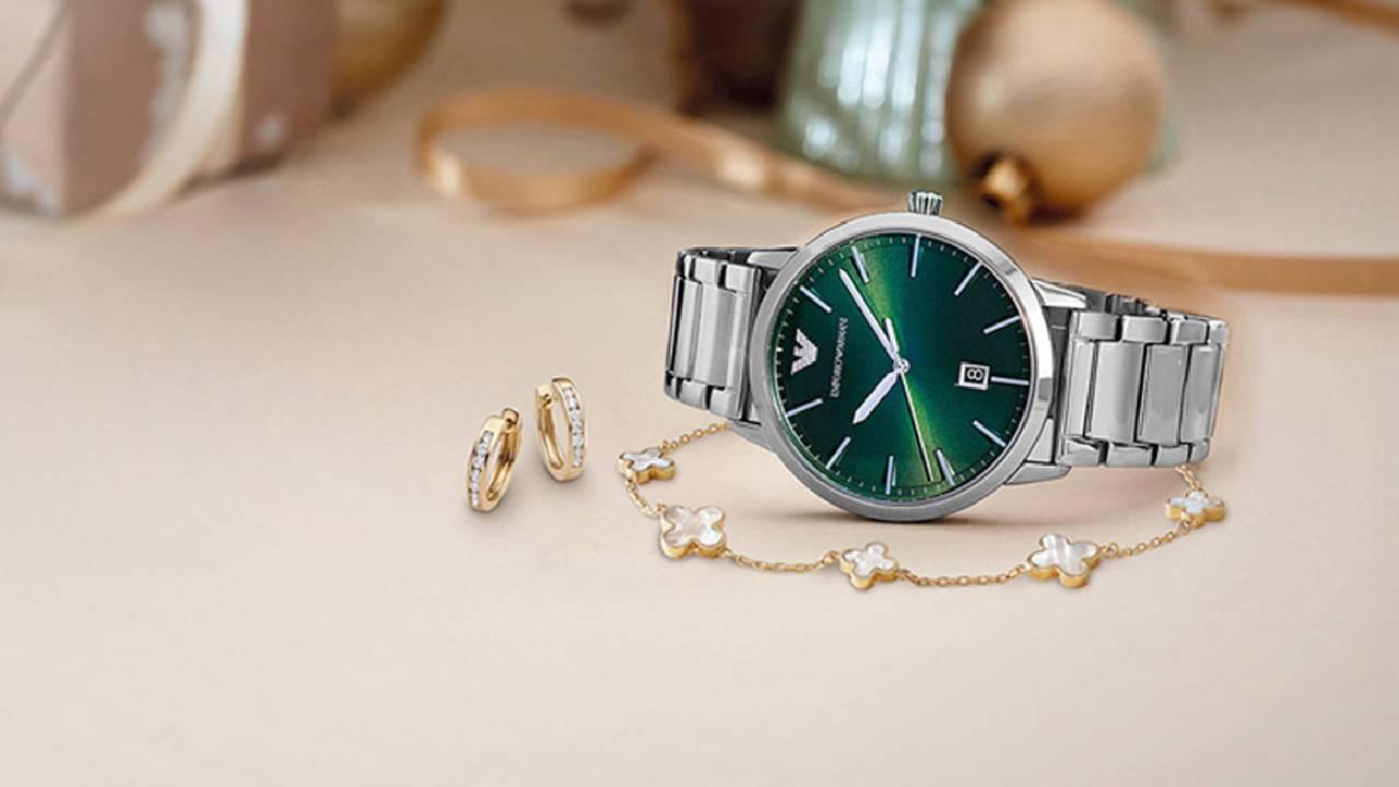  A silver watch with a emerald green watch face with a gold bracelet and a pair of gold earrings from Beaverbrooks in front of a festive backdrop. 