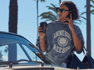 A woman on her phone holding a coffee cup next to a car with palm trees in the background wearing a shirt from the Anine Bing x Reebok collab