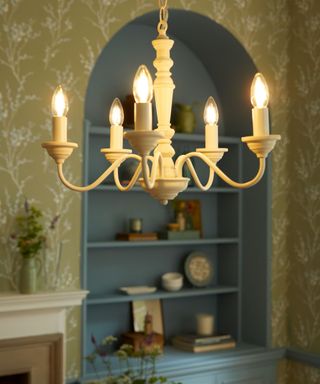 A white six candle chandelier hanging from a ceiling with yellow patterned wallpaper and a light blue arched bookcase with decor on it behind