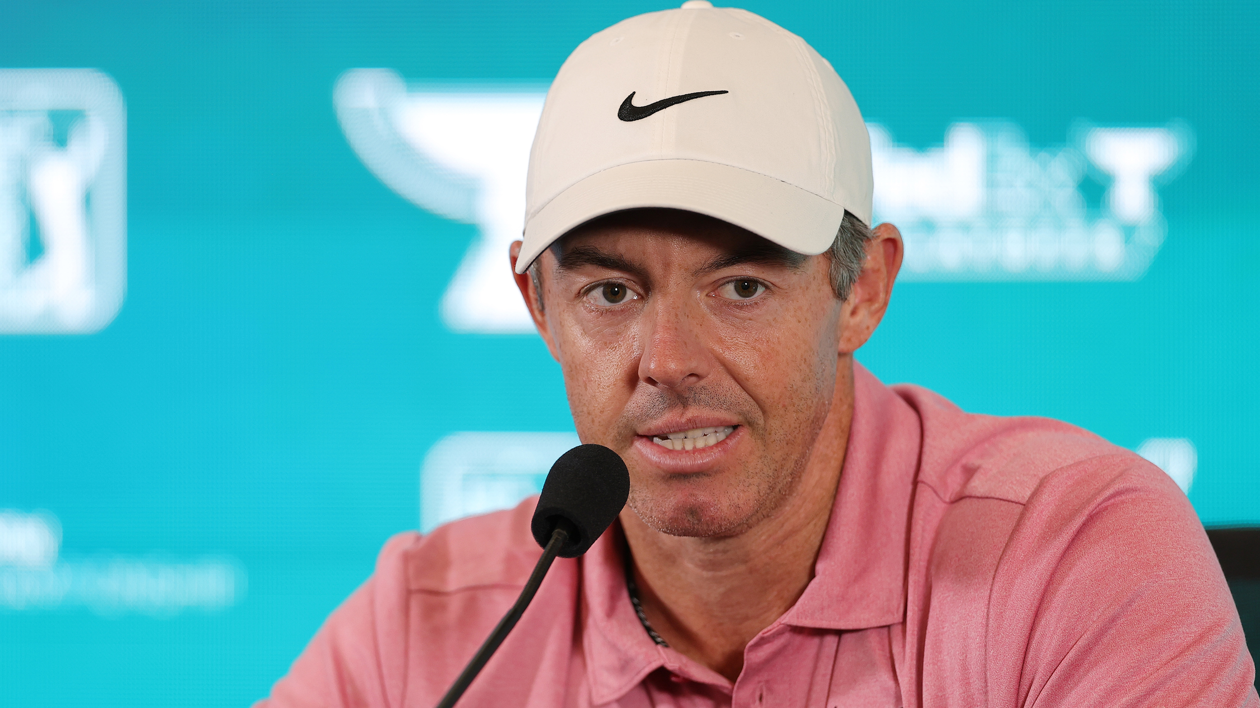 Rory McIlroy wearing a pink polo and white cap speaking in front of a blue sponsor background at his BMW Championship press conference