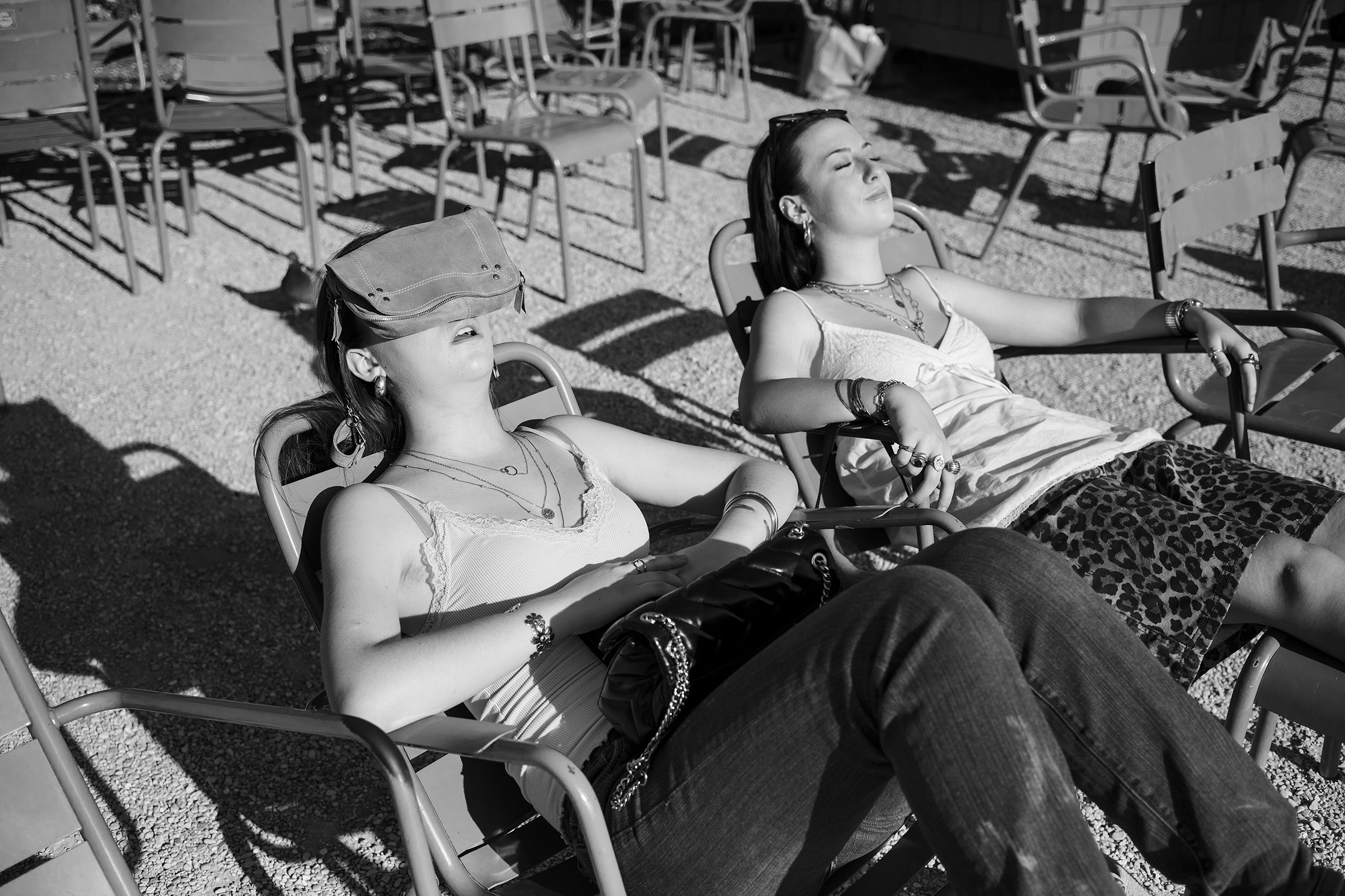 Black-and-white scene of two women reclining in patio chairs among empty seats, one shielding her eyes with a cap as the other lounges in the sun, relaxed mood