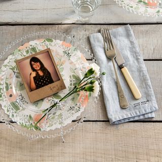 A rustic dining table with a glass and floral plate with a polaroid picture used as a place setting