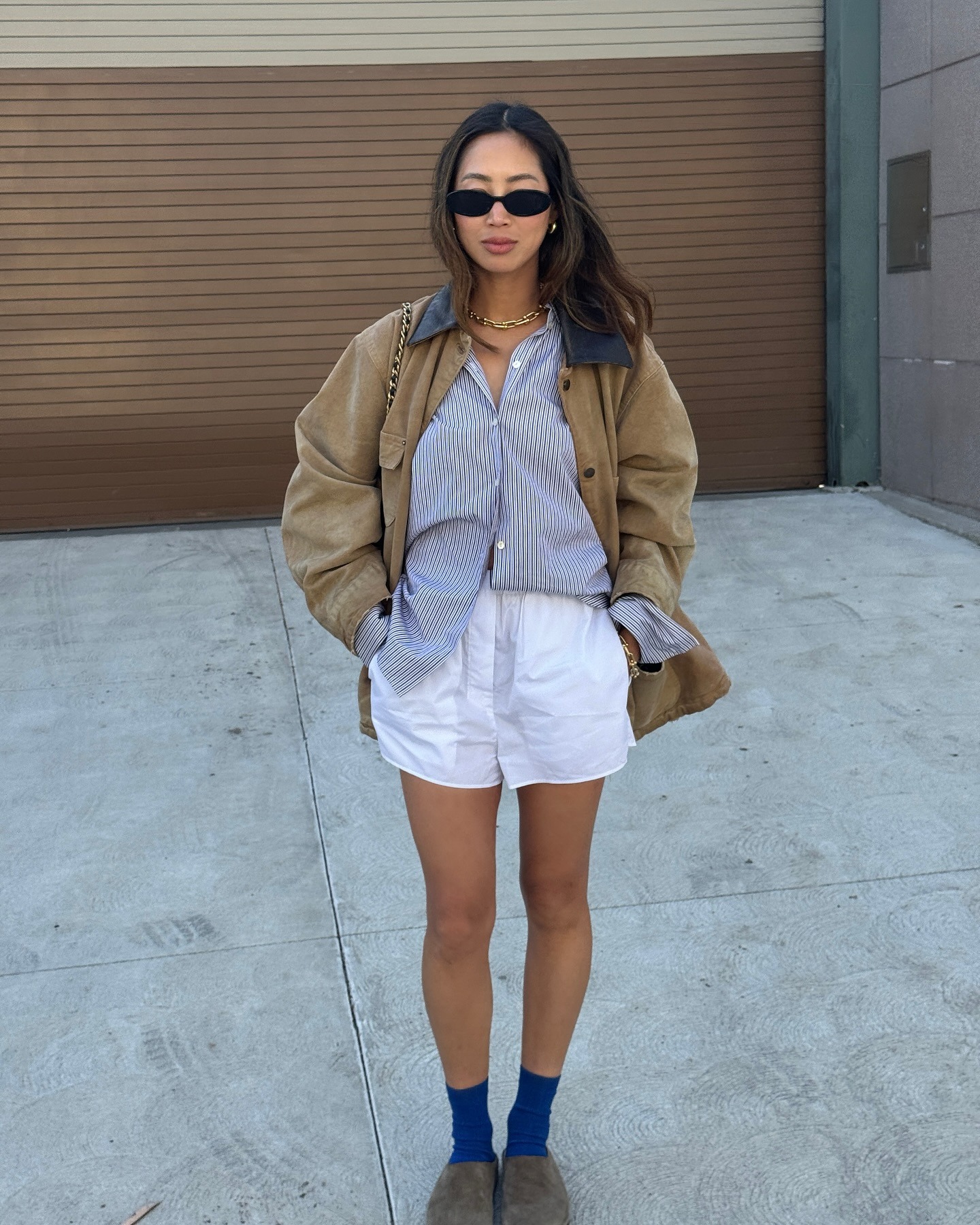 Woman wears Comme Si blue socks, suede clogs, white boxer shorts, striped button dow and workman jacket.