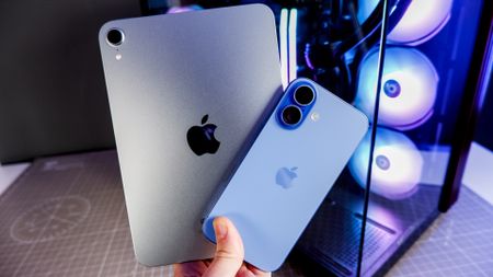 An iPad Mini A17 and iPhone 16 on a desk with a gaming PC in the background.