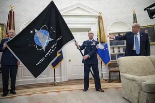 A bald man in a dark formal military uniform holds a flag pole with a large space force flag in a white room. To the right, a fat, overgrown baby holds his hand out in confusion.