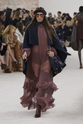 A model at Chloé's Fall/Winter 2024 show wearing a sheer, ruffled mauve maxi dress, a navy cape, a large clutch, thigh-high boots and pink sunglasses.