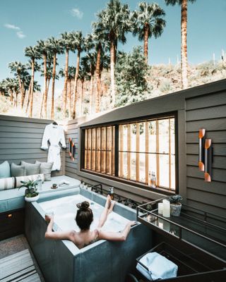 A luxury decked backyard space with outdoor hot tub and seating area with palm style trees and adjacent outbuilding