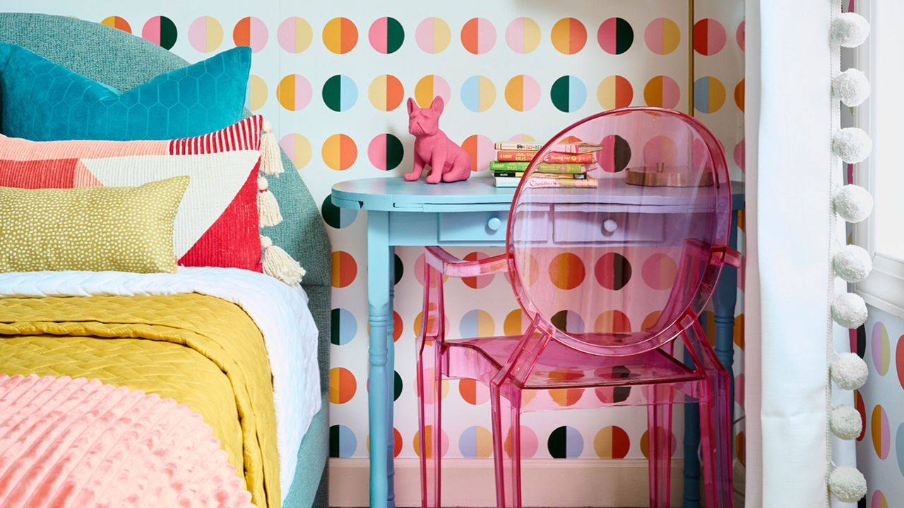 bedroom filled with joyful colors. transparent pink chair at blue desk, patterned wallpaper behind a blue bedhead with yellow, pink and red sheets