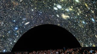 An audience looks at an arced screen dotted with stars and galaxies as the first images from the Rubin Observatory are revealed. 