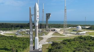 a white rocket is rolled toward a launch pad with greenery along the sides and the ocean in the background.