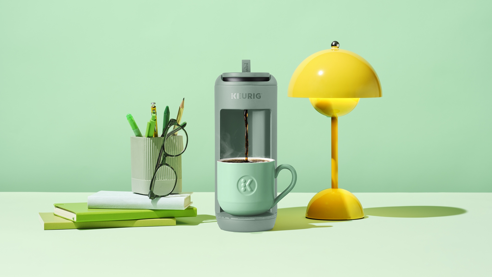 The Keurig K-Mini Mate in Glamping Green with a lamp, glasses and books beside it