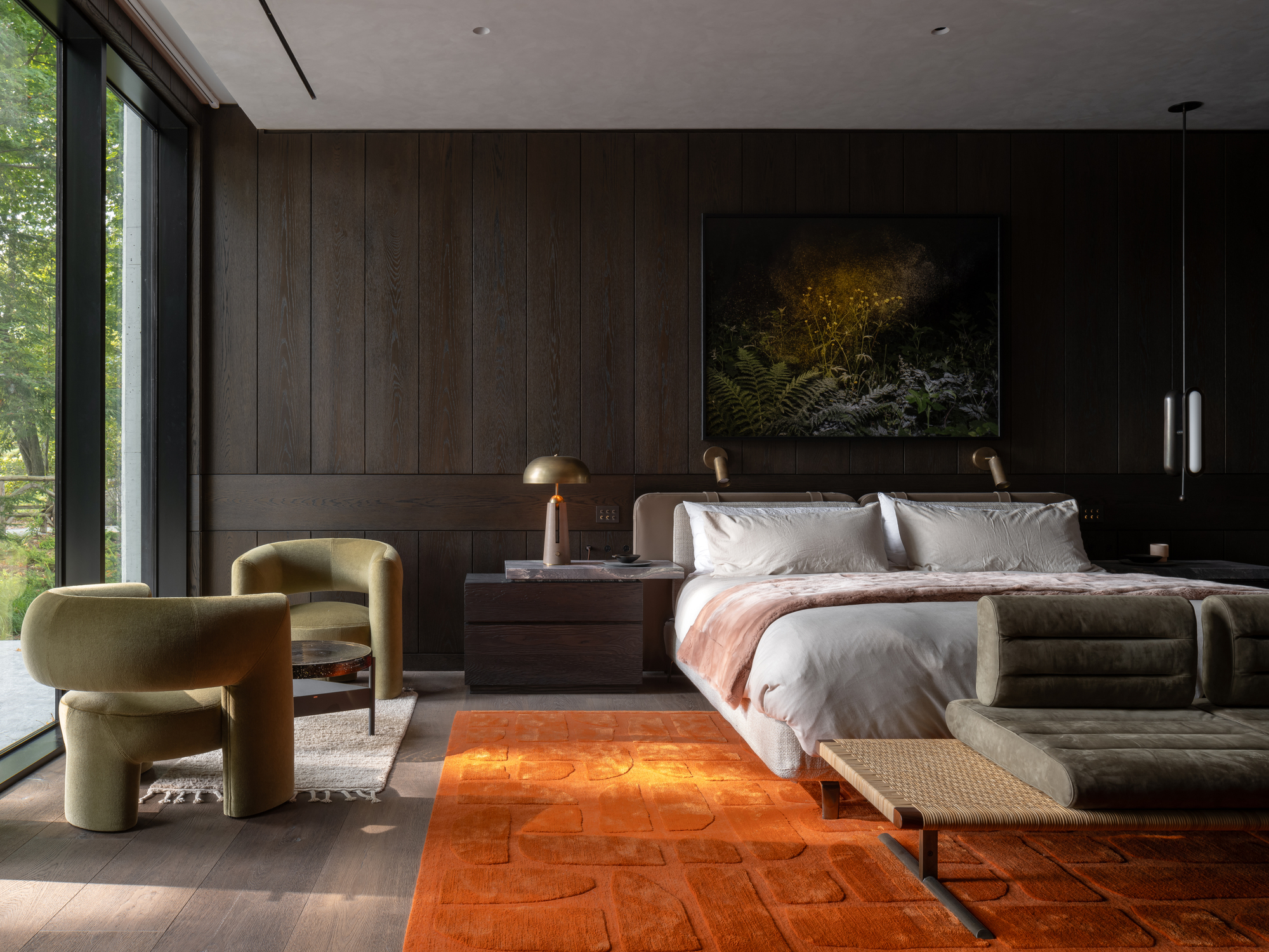 modern bedroom with dark timber paneling on the walls, orange carpet, gray bedding, bench seat at end of the bed, gold mushroom lamps on bedside table, and two green velvet armchairs by the window