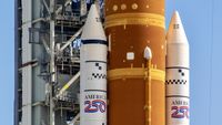 The mid section of an orange rocket with white side boosters stands against a metal tower. America 250 is decaled on both boosters.