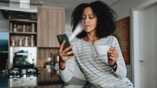 Person, sat in a kitchen holding a mug, scanning their face with their smartphone