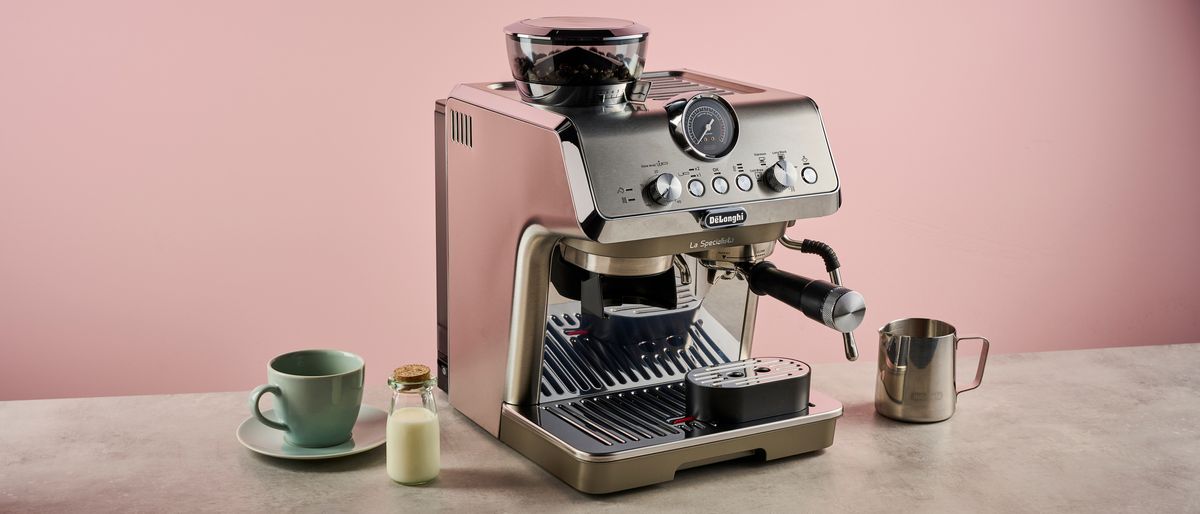 The De&#039;Longhi Specialista Arte Evo sits on a stone-effect surface in front of a pink backdrop. It has a green cup and saucer, and a small cork-topped bottle of milk to its left, and the included metal milk jug on its right.