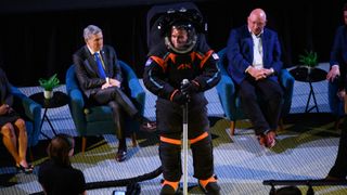 an engineer demonstrates a black and orange Axiom Space spacesuit on a stage
