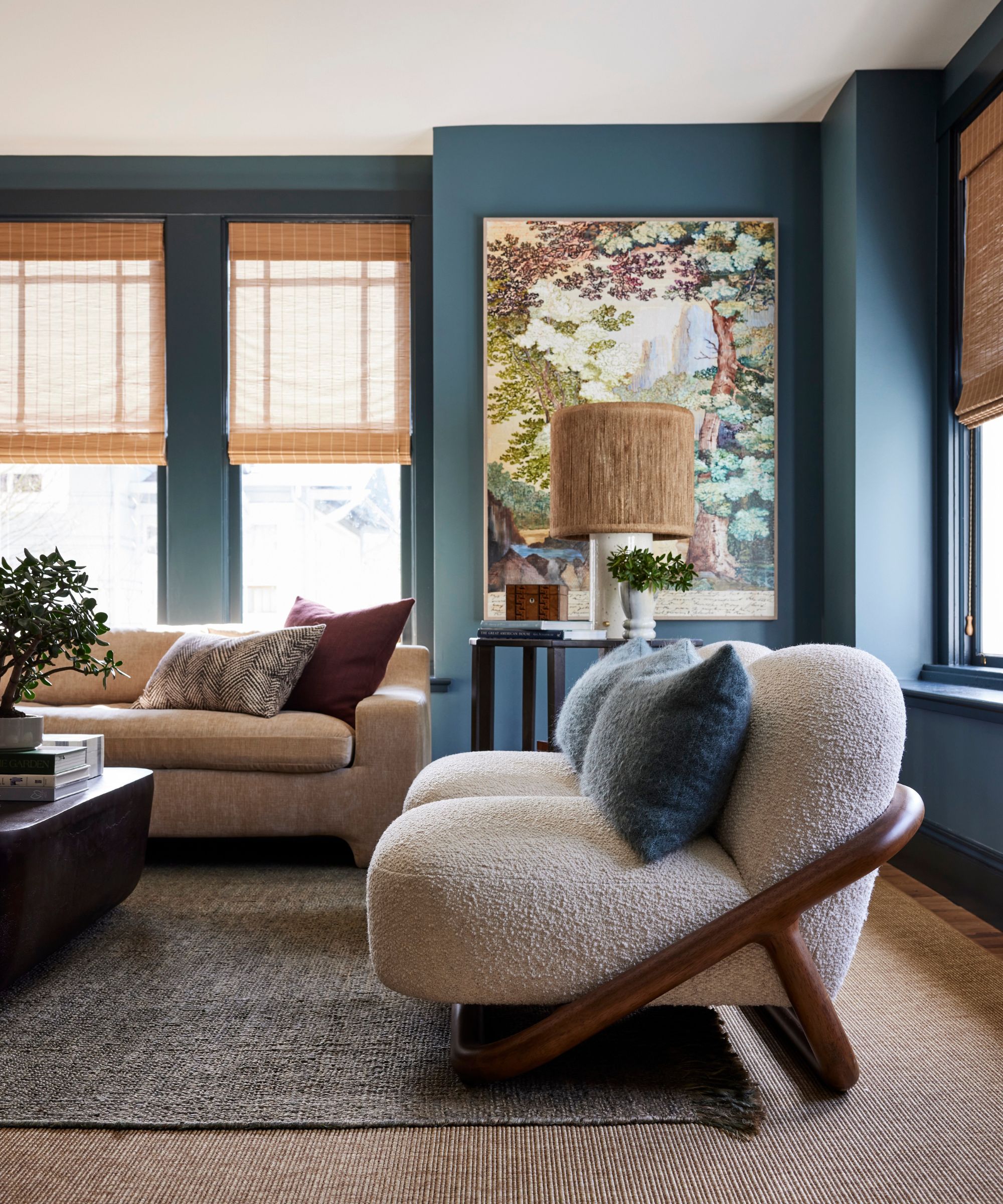 A living room with blue-gray walls, a beige carpet and a gray rug, a cream boucle sofa and a beige sofa in front of windows with woven roller blinds.