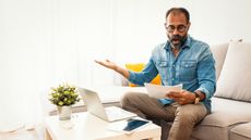 A frustrated-looking man looks at paperwork while sitting on his sofa.