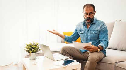 A frustrated-looking man looks at paperwork while sitting on his sofa.