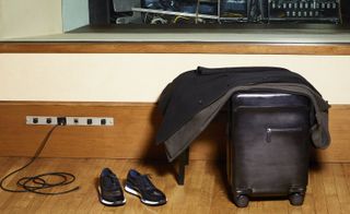 A black leather suitcase on wheels with a jacket on top of it and shoes next to it in front of a wooden skirting board with a row of plugs on it.