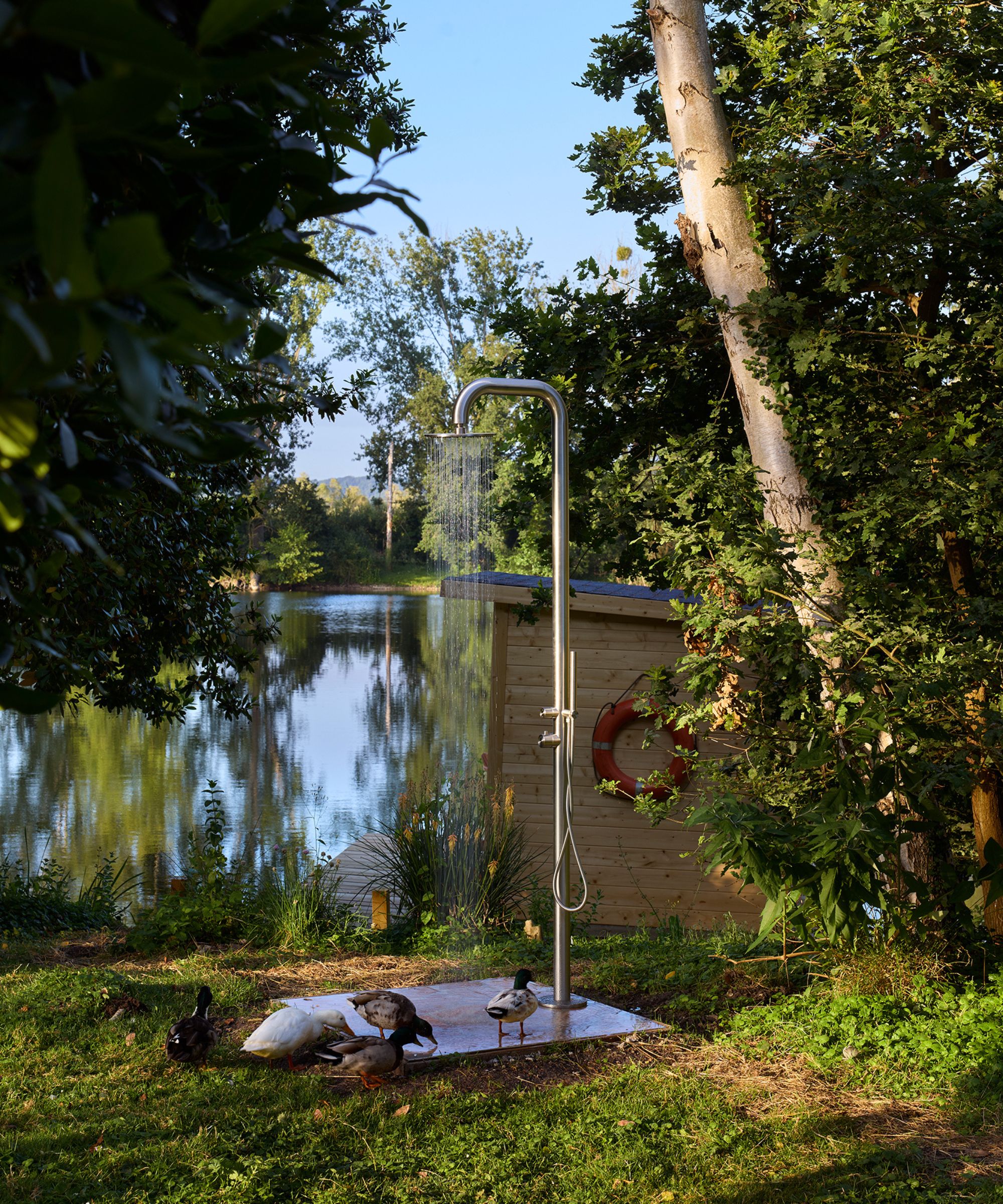 an outdoor shower set by a lake in France with a small boat house and ducks