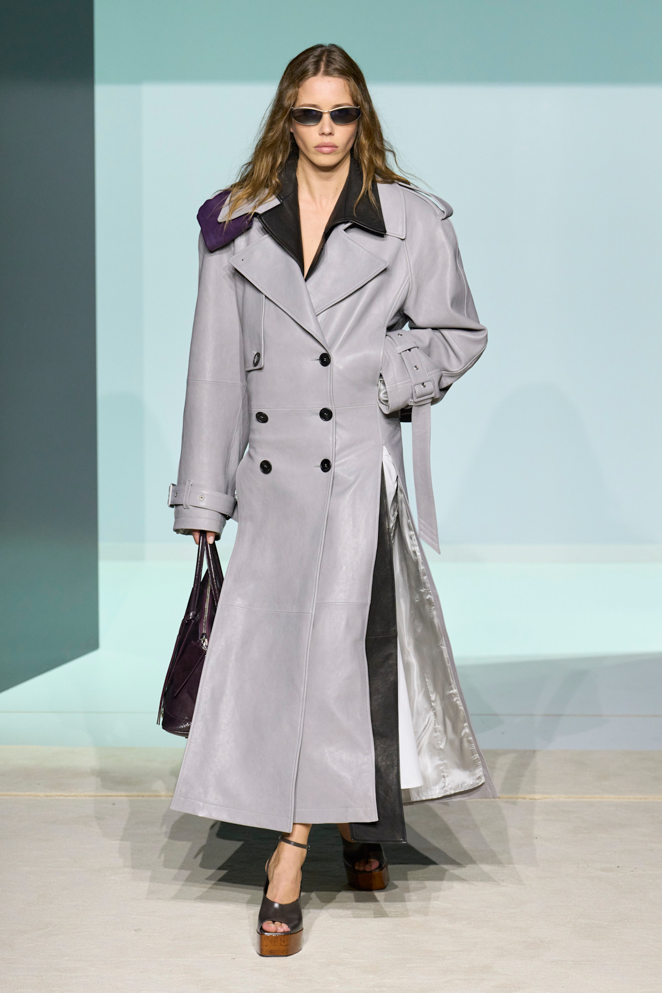 Layering staples for spring 2026 are shown in a photo from The Attico's runway show, with a model walking while wearing a gray leather trench coat layered over a black trench coat styled with black and brown peep toe platform heels, a black handbag, and black skinny sunglasses 