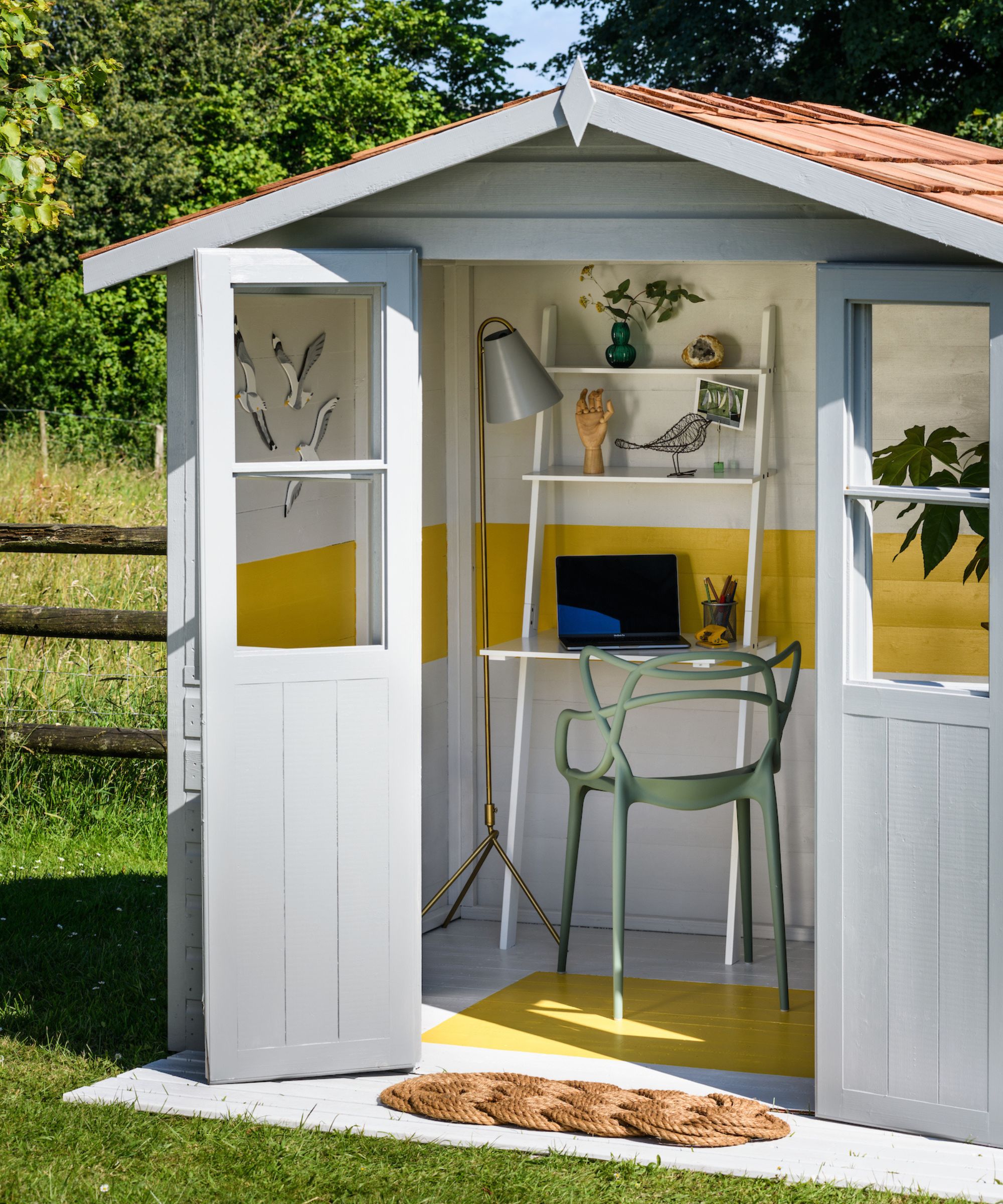 A grey painted garden shed converted into a home office, open to reveal yellow painted interior walls and floor, a ladder desk, green chair and brass floor lamp