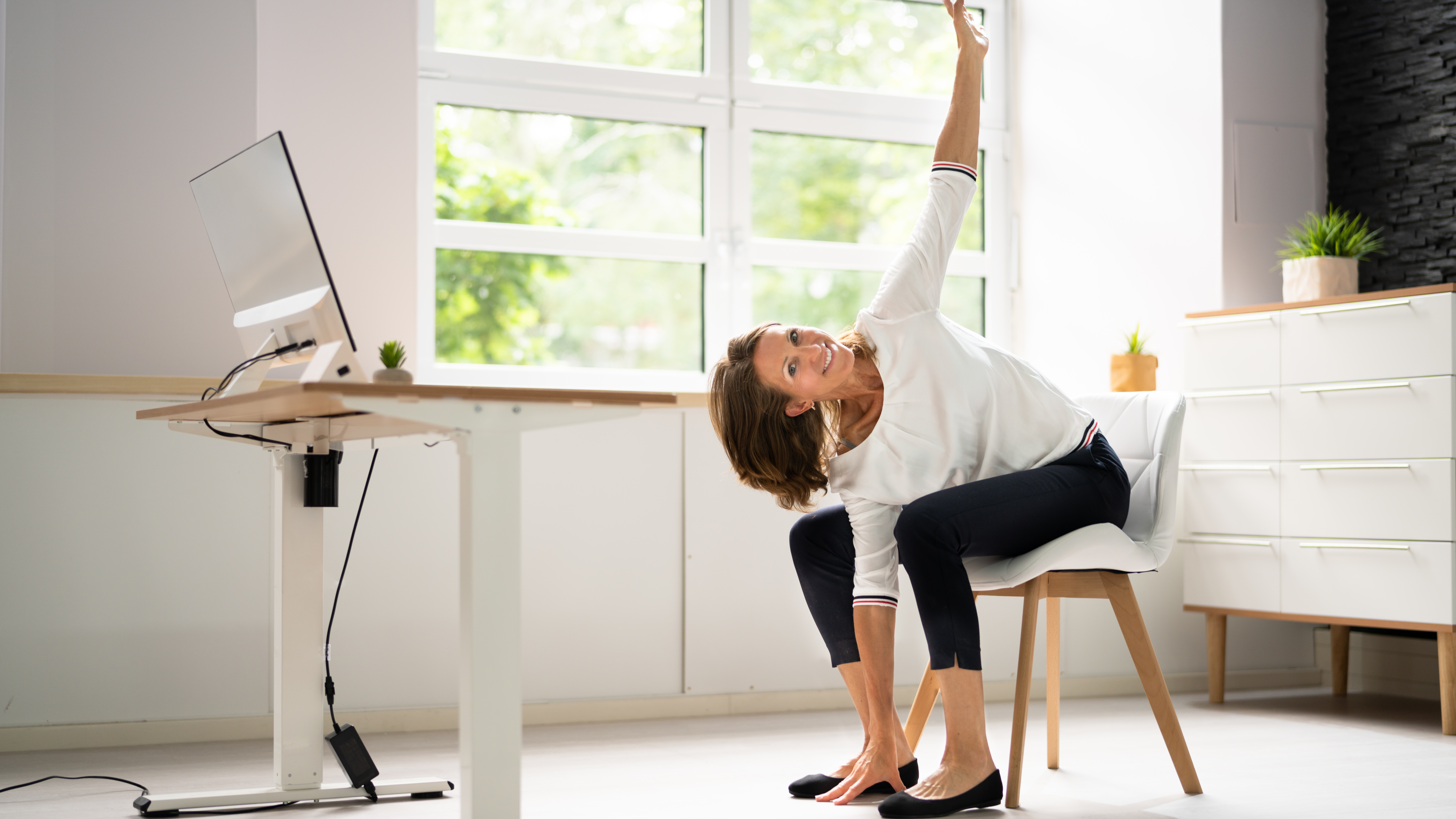 A woman in officewear sits in a chair performing a seated windmill. Her legs are hip-width apart, feet on the floor, her torso is twisted toward her left leg and her right hand is touching the floor as her left hand reaches toward the ceiling. Her torso 