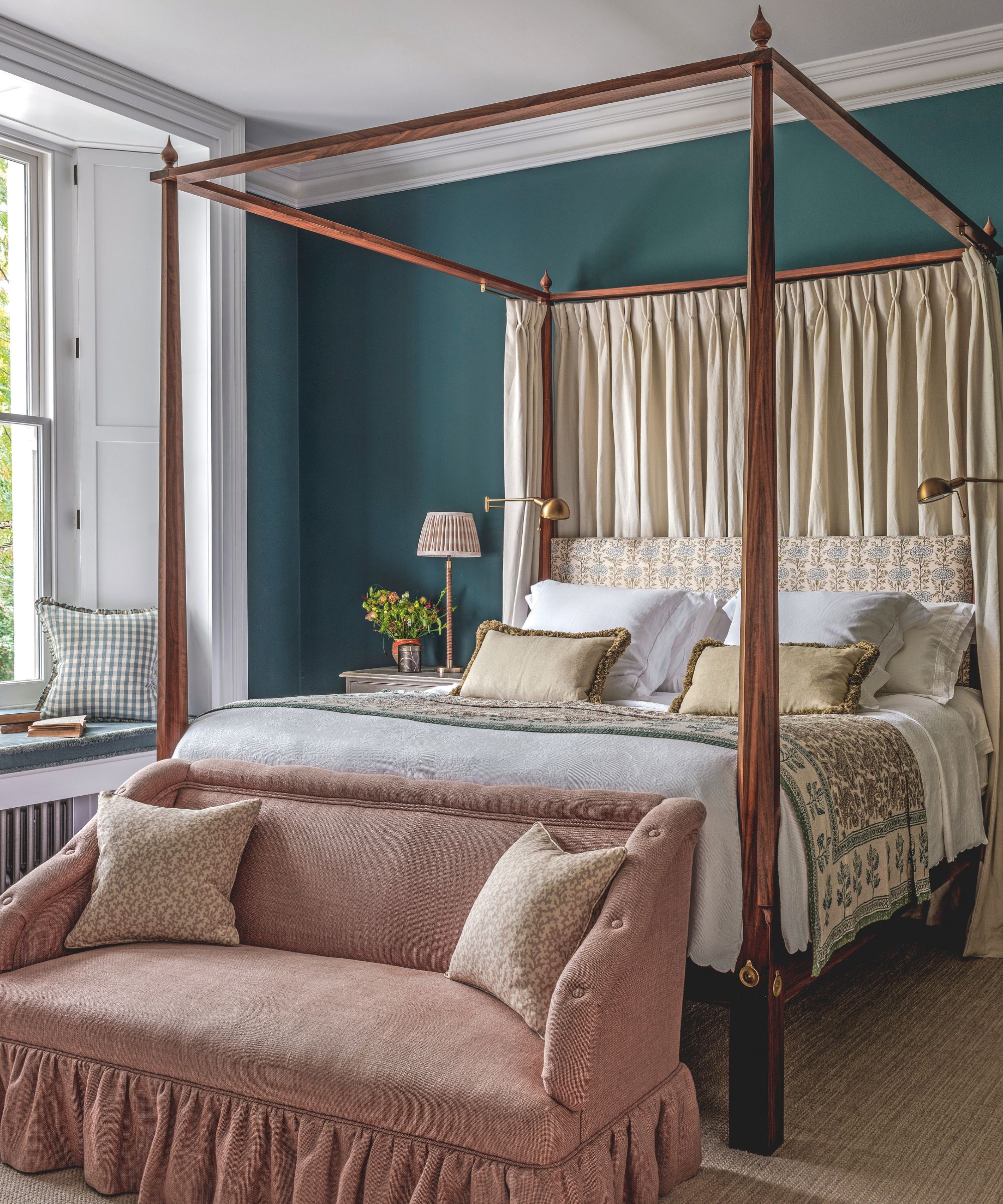 A traditional bedroom featuring a tall wooden four-poster bed with a teal accent wall, white bedding, and a dusty rose upholstered bench at the foot.