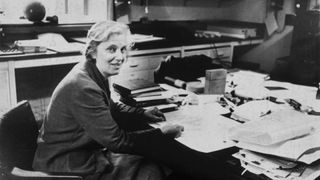 Black and white photograph of Professor Dorothy Mary Hodgkin taken in the 1960s. She is sitting at a large desk overflowing with papers and books.