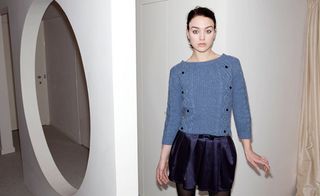 Female model, wearing blue knitted jumper, dark blue short skirt and tights, white walls, large white framed oval mirror