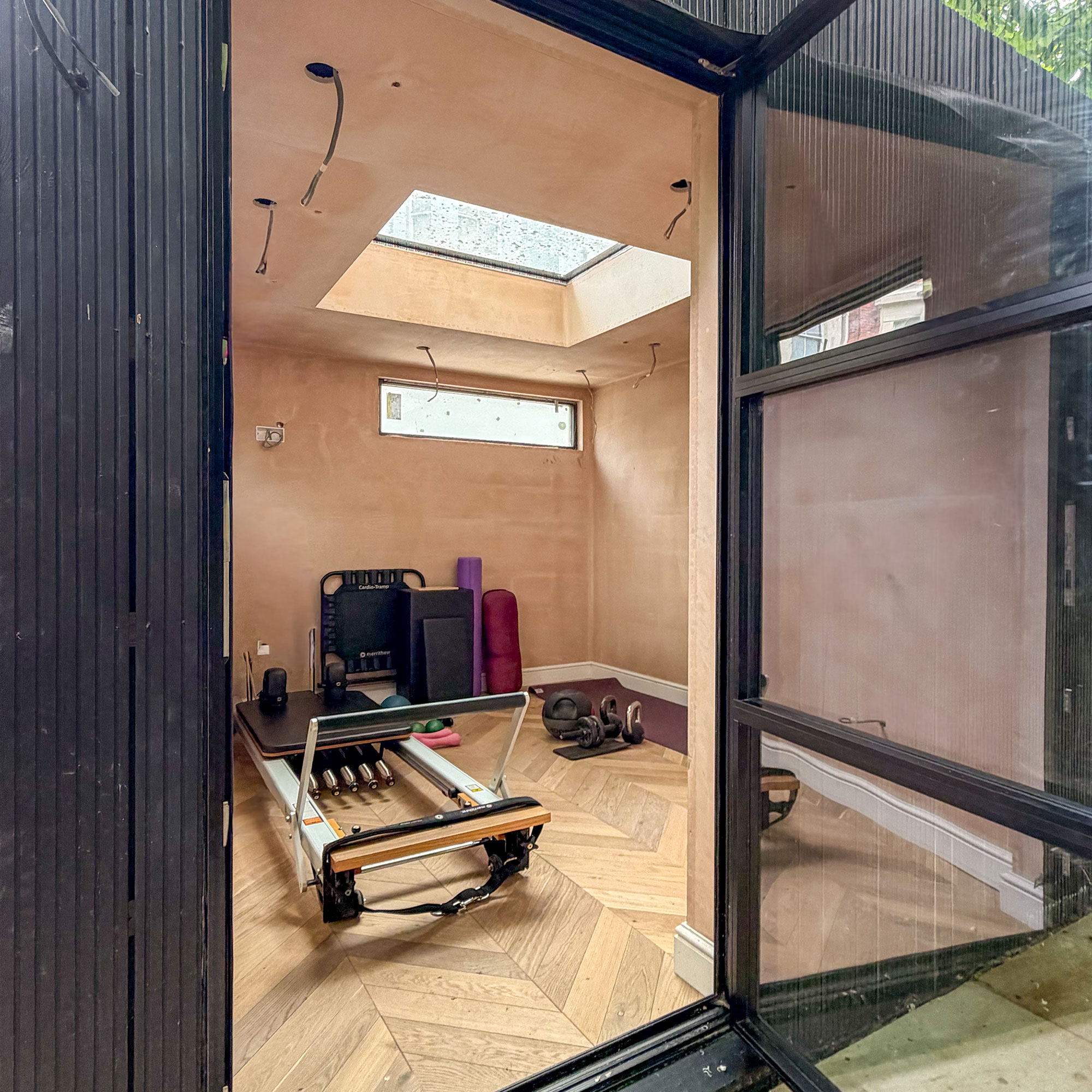 Home gym mid renovation with plaster walls, exposed wiring, pilates reformer machine and wooden parquet flooring