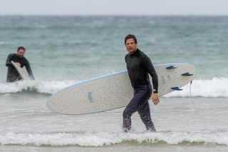 Pete (James Norton) stands at the edge of the sea in a wetsuit with a surfboard under his right arm as he steps towards the beach. In the background, Miles (James McArdle) is also in a wetsuit and picking up his surfboard.