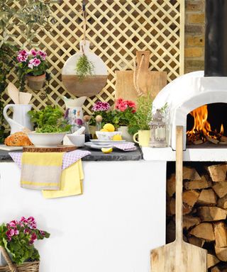 A white outdoor kitchen with pizza oven, white crockery, yellow accents and a wooden trellis backing.