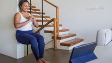 A woman performs a wall squat at home next to a stair case. Her back is against the wall, her knees are bent and her hands clasped in front of her. She is smiling at a laptop in front of her.