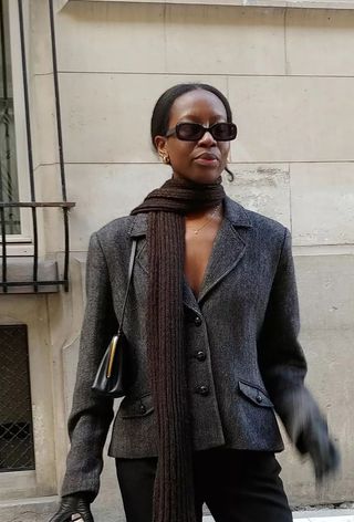 A woman wearing a tailored gray blazer with a brown scarf, a black Frame bag, black leather gloves, black sunglasses, and gold earrings.