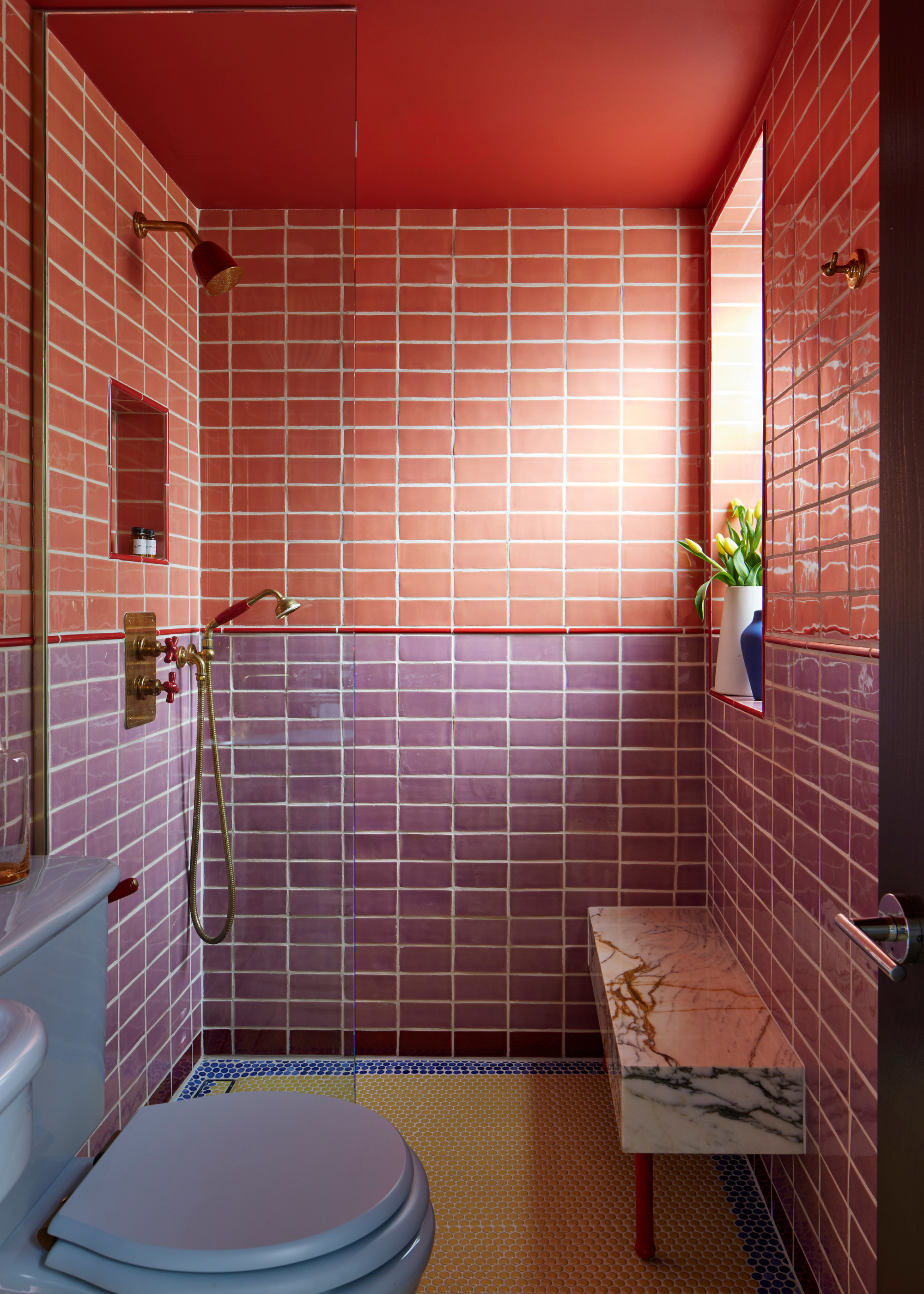 A colorful small bathroom with glossy purple and peach tiles, a marble shower bench, and a blue toilet
