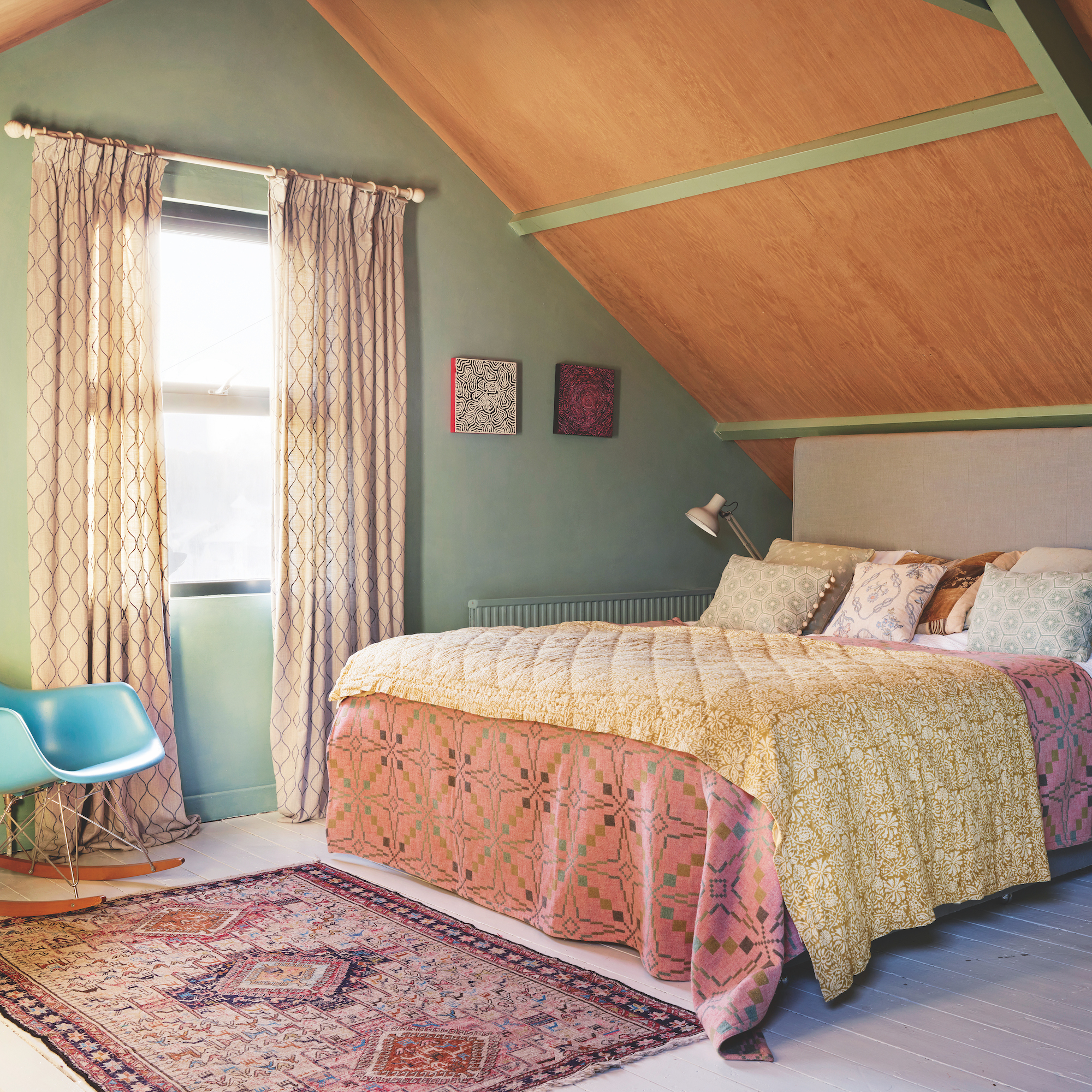 eaves bedroom with sloping wood panel roof above bed dressed with quilt and Welsh blanket, rug on floor and eames rocking chair