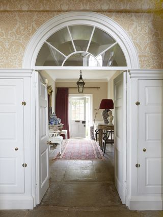 grand entryway to period property, wallpaper, arched window, stone flags, entryway closets on each side, console tables, front door in background, pendant light, table lamps