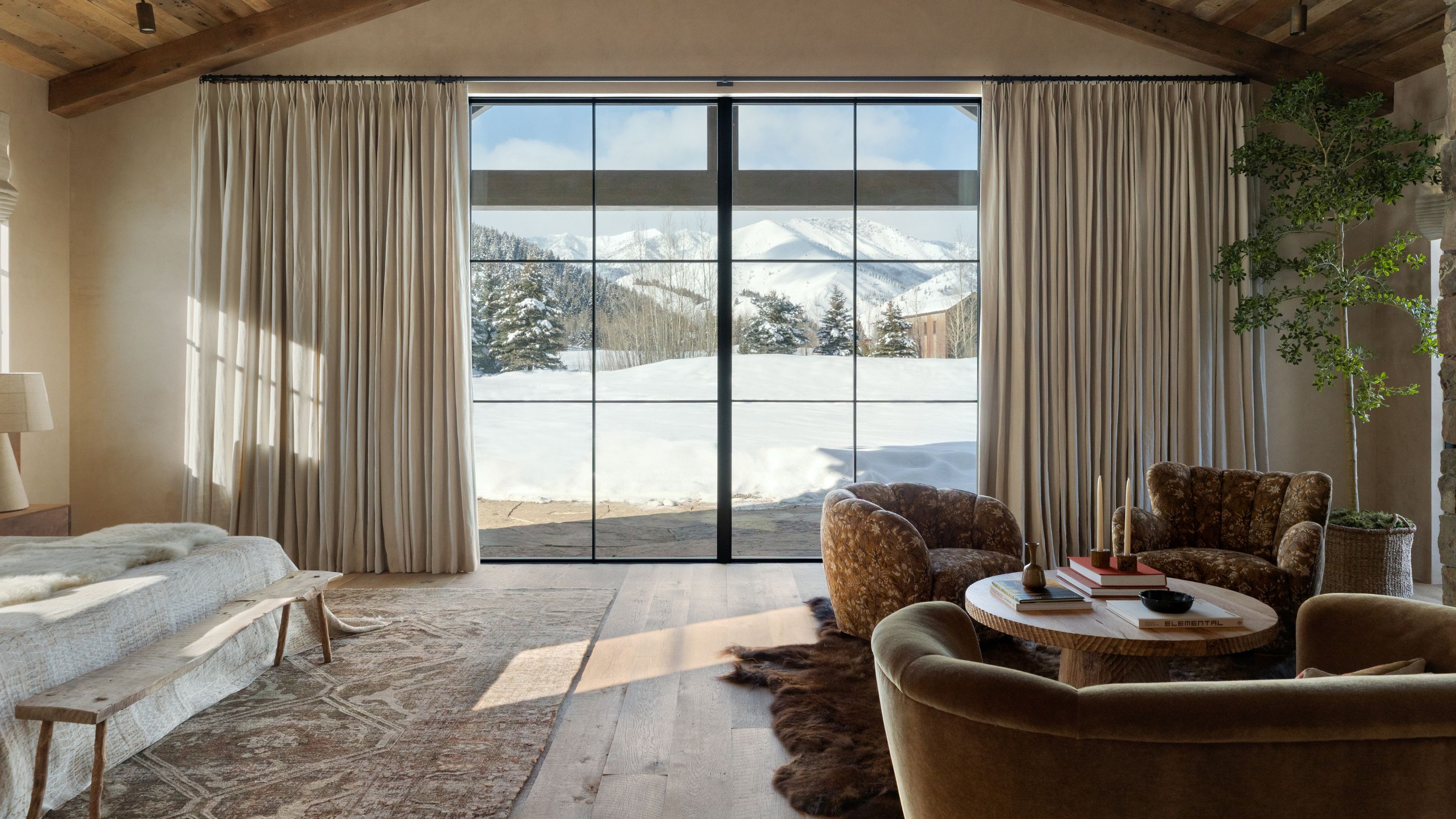 a large primary bedroom with vast glass doors framing a snowy mountain view, vaulted ceiling, a neutral linen dressed bed with a seating area on the right