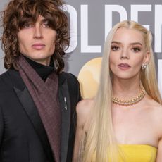 Anya Taylor-Joy wears a strapless yellow top and matching skirt and has long blonde hair while her husband Malcolm McRae wears a dark suit jacket with a brown scarf