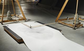 A drawing machine. A square board on the floor with a long sheet of white paper and 2 pendulars at 90 degrees angle