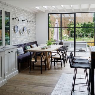 white colored room with a wooden and patterned tile floor, with a wooden dining table and a purple sofa with patterned cushions and black chairs with a beige woven sitting-top next to glass black paneled doors and a garden