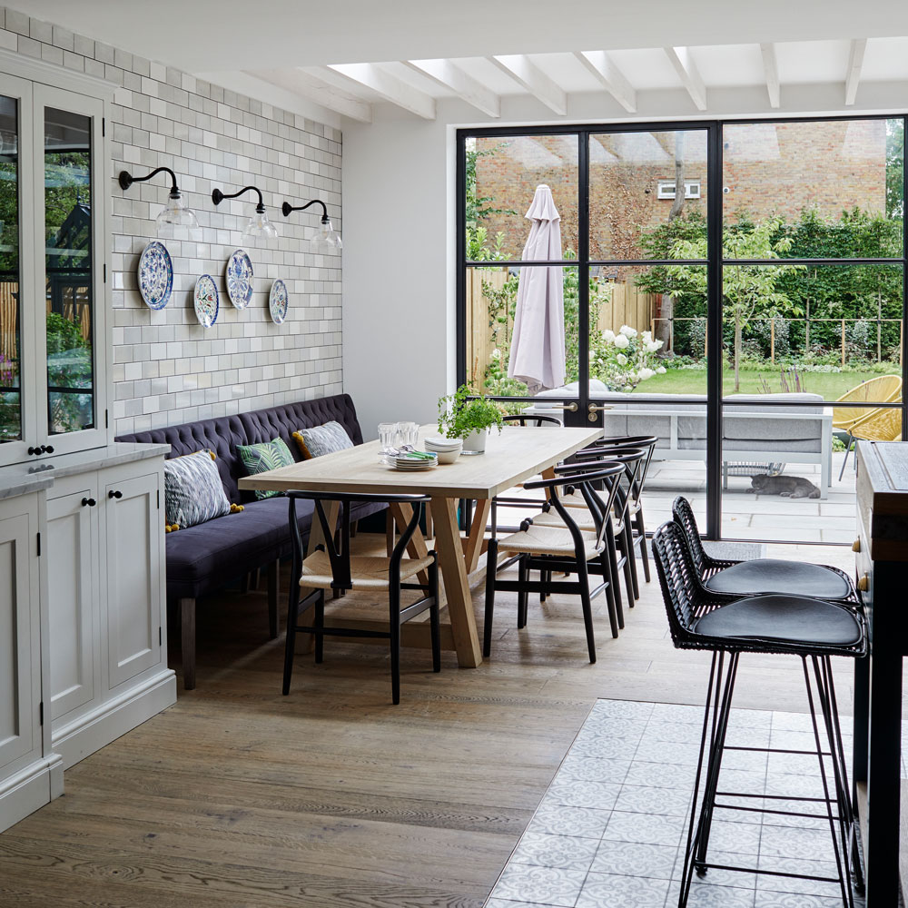 white colored room with a wooden and patterned tile floor, with a wooden dining table and a purple sofa with patterned cushions and black chairs with a beige woven sitting-top next to glass black paneled doors and a garden
