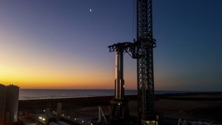a big silver rocket stands on a launch pad at dawn or dusk, with the ocean in the background
