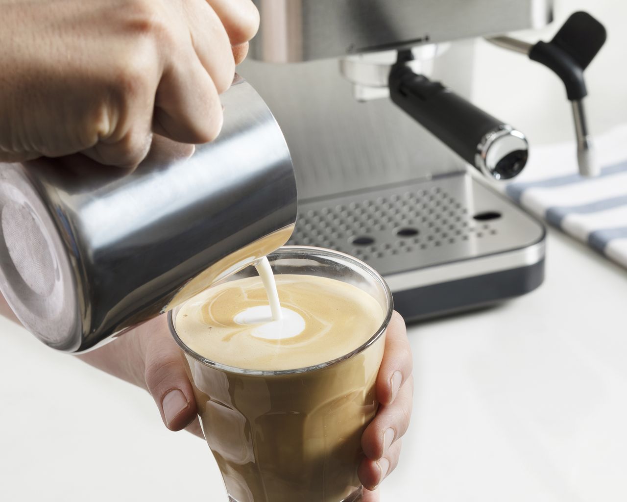 A person using a Breville Bijou coffee maker and metal milk jug to pour milk into glass cup in order to create latte coffee art