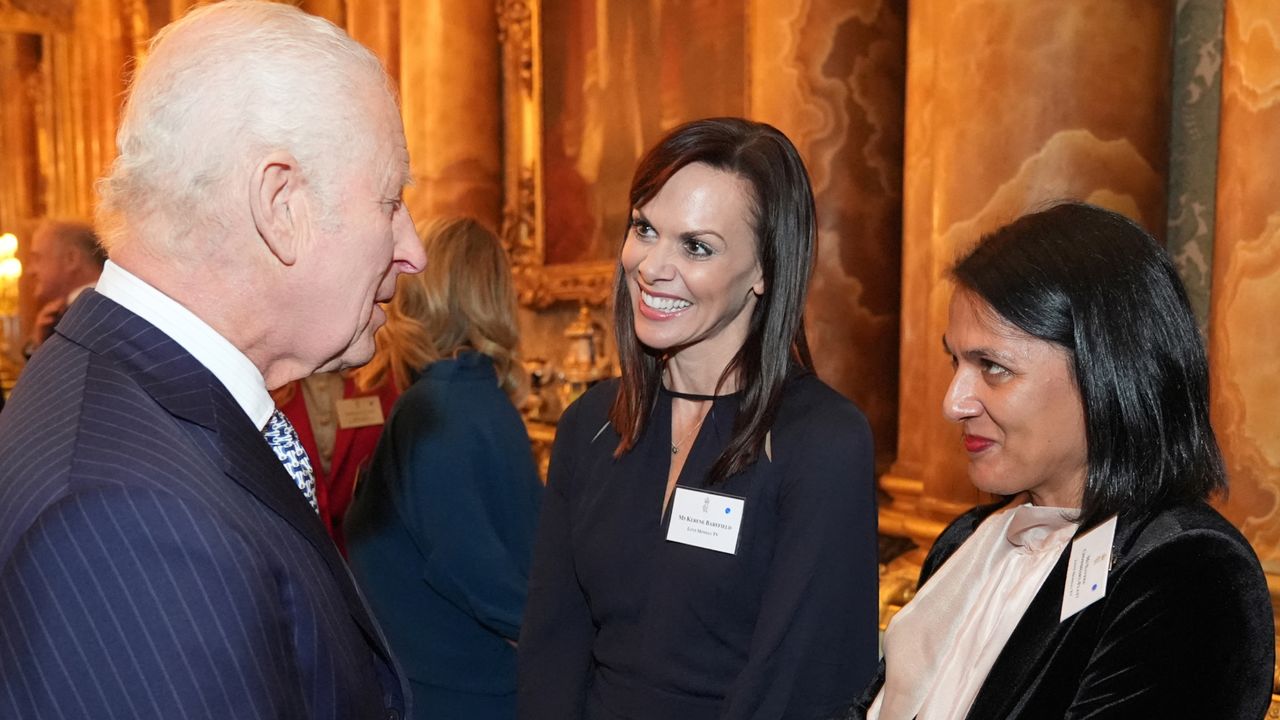 King Charles wearing a blue suit while speaking to filmmakers at a Buckingham Palace reception and shaking hands with Naveed Chowdhary-Flatt who is wearing a black velvet blazer and cream pussybow blouse